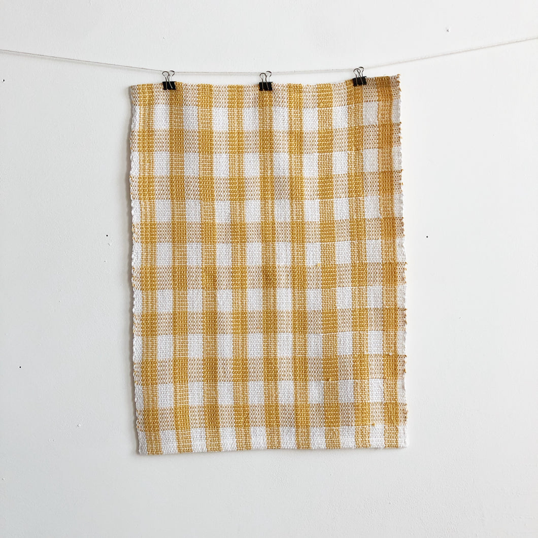 a handwoven white and yellow checked teatowel hangs on a clothesline in front of a white wall
