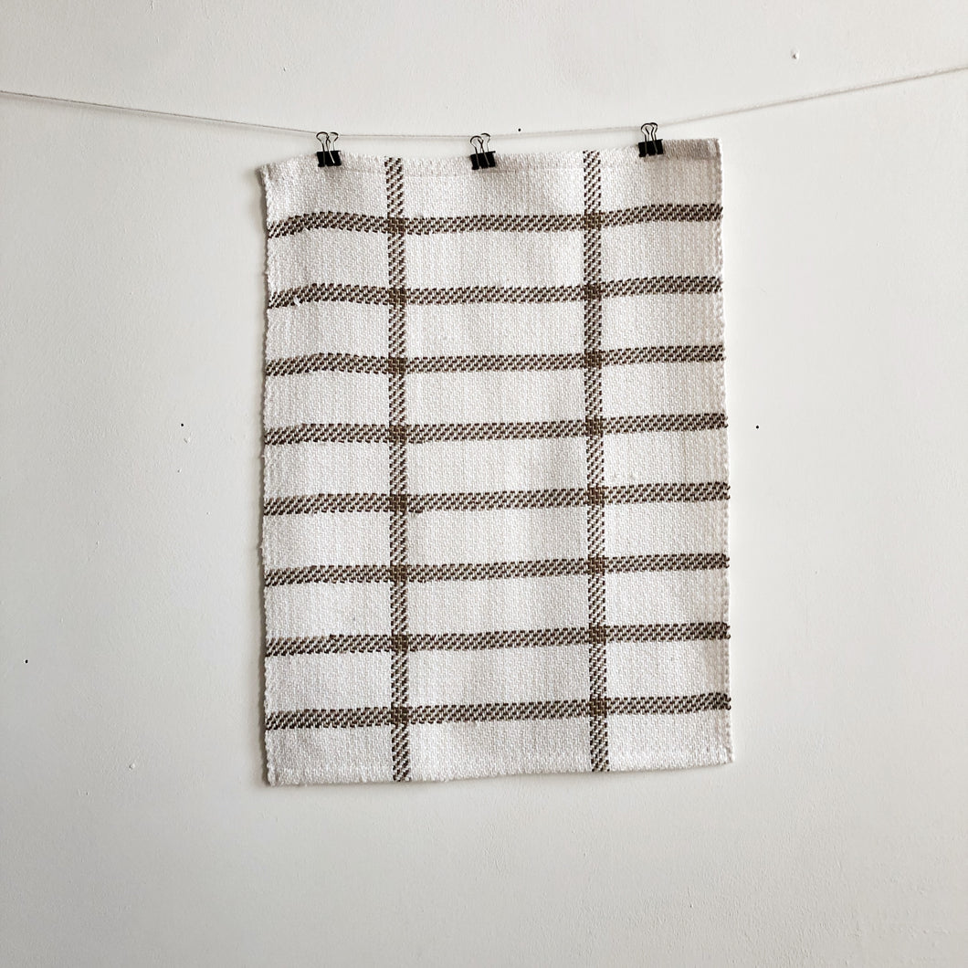 a handwoven white and brown teatowel hangs on a clothesline in front of a white wall