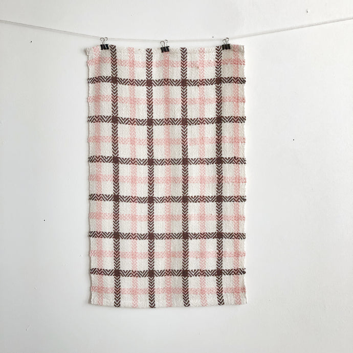 a handwoven white, pink and brown teatowel hangs on a clothesline in front of a white wall