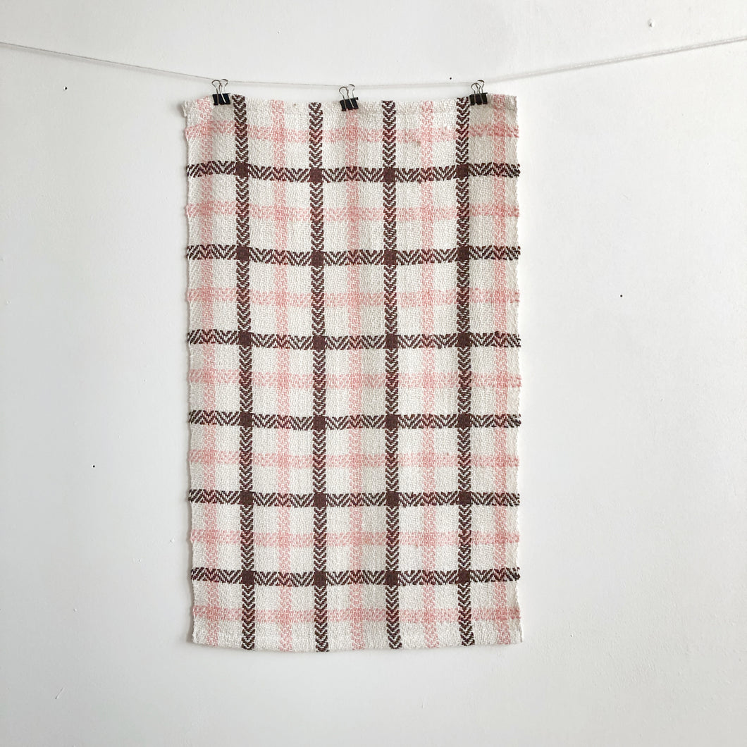 a handwoven white, pink and brown teatowel hangs on a clothesline in front of a white wall