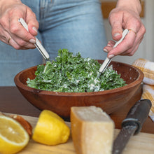 Load image into Gallery viewer, a woman&#39;s hands toss a green salad in a wooden bowl on a table surrounded by squeezed lemons, a block of cheese and a white and yellow tea towel
