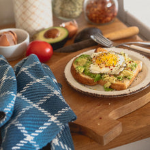 Load image into Gallery viewer, a slice of avocado toast with a fried egg on top sits on a white plate on a wood counter surrounded by tomatoes, half an avocado, egg shells and a blue checked teatowel
