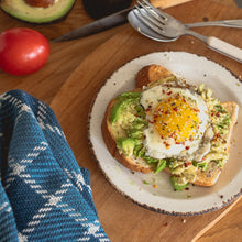 Load image into Gallery viewer, a slice of avocado toast with a fried egg on top sits on a white plate on a wood counter surrounded by a tomato, half an avocado and a blue checked teatowel

