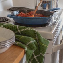 Load image into Gallery viewer, a stack of white bowls and a green teatowel sit on the counter next to a blue pan filled with red sauce being stirred on the stove
