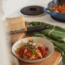 Load image into Gallery viewer, a bowl of pasta in red sauce garnished with cheese and fresh basil sits next to a green teatowel on the counter next to a stove with a blue pan on it
