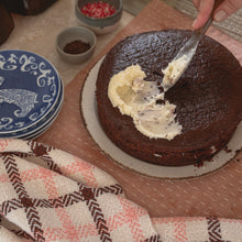 Load image into Gallery viewer, a close up of a chocolate cake being iced with white icing next to a stack of blue plates, small bowls of spinkles and a white, pink and brown teatowel
