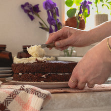 Load image into Gallery viewer, a woman&#39;s hands spread white icing on a chocolate cake next to a white, pink and brown tea towel. there are purple irises in the background
