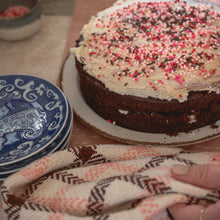 Load image into Gallery viewer, a chocolate cake with white icing and pink sprinkles sits on the counter next to a stack of blue plates and a white, pink and brown teatowel
