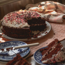 Load image into Gallery viewer, a chocolate cake with white icing and pink sprinkles sits next to two cake slices on blue plates with a pink, white and brown teatowel in the background
