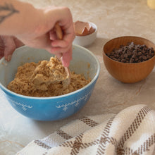 Load image into Gallery viewer, a woman&#39;s hand stirs cookie dough with a wooden spoon in a blue bowl next to a a cracked egg, a bowl of chocolate chips and a white and brown teatowel on the counter
