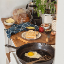 Load image into Gallery viewer, an egg fries in a cast iron pan next to a messy counter with a slice of toast, egg shells, tomato, avocado and a blue checked tea towel

